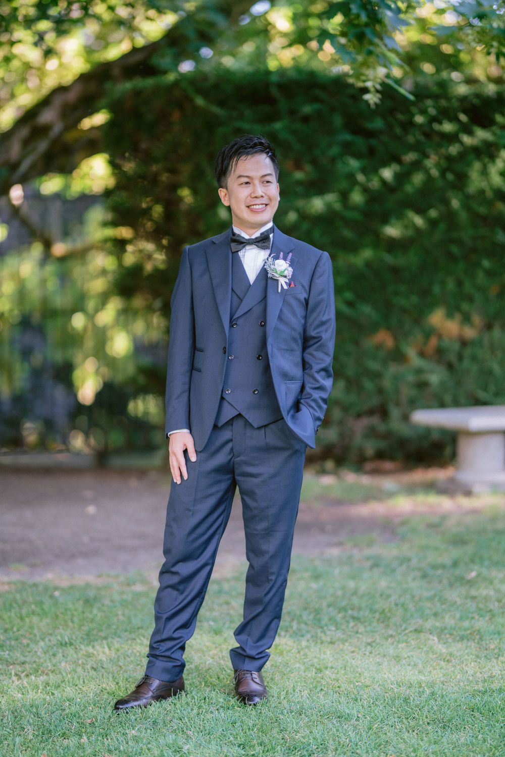 Groom in navy three-piece suit poses in a lush garden setting for wedding portraits.
