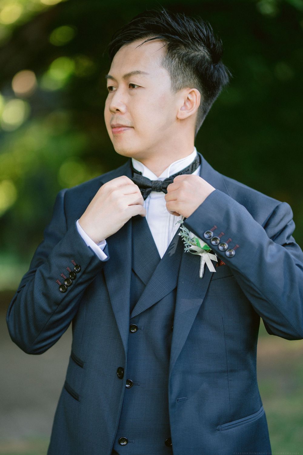 Groom adjusting bowtie while wearing navy suit with floral boutonniere.