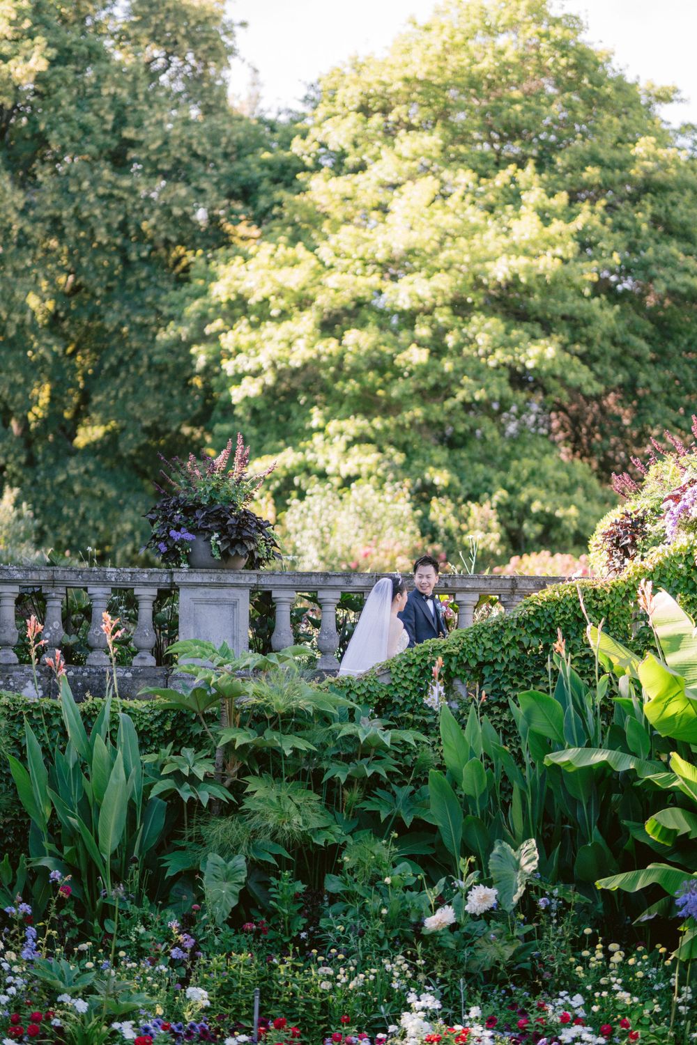 Wide garden landscape with flowering plants and couple visible in background among stone architecture.