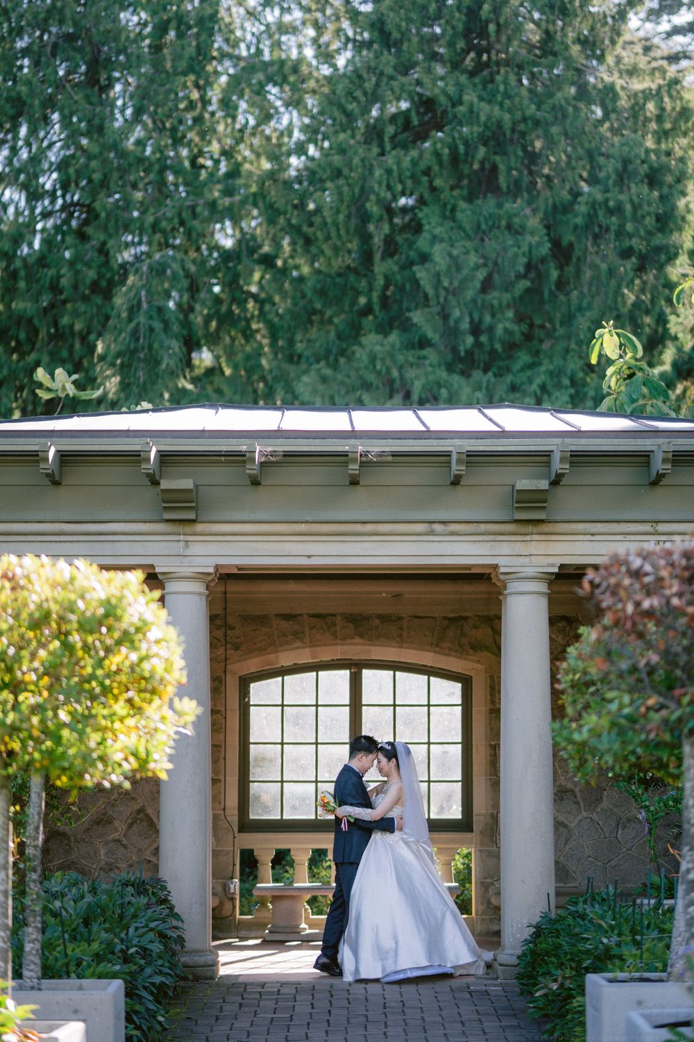 Wedding couple poses romantically under an elegant garden archway with white columns and decorative roof.