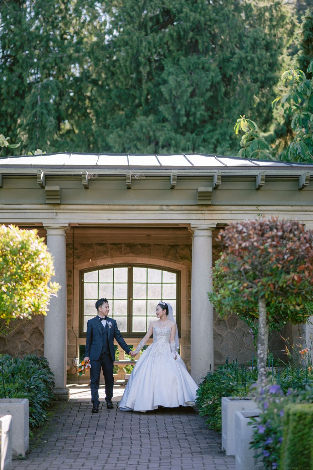Elegant couple stands beneath a classical stone portico in a lush garden setting.