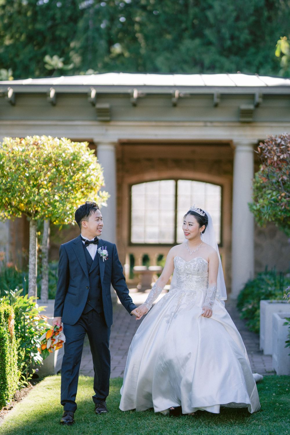 Couple walks through a manicured garden path beside an ornate archway.