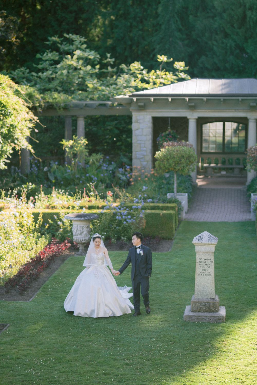 Couple poses on manicured lawn near stone monument and classical architecture.