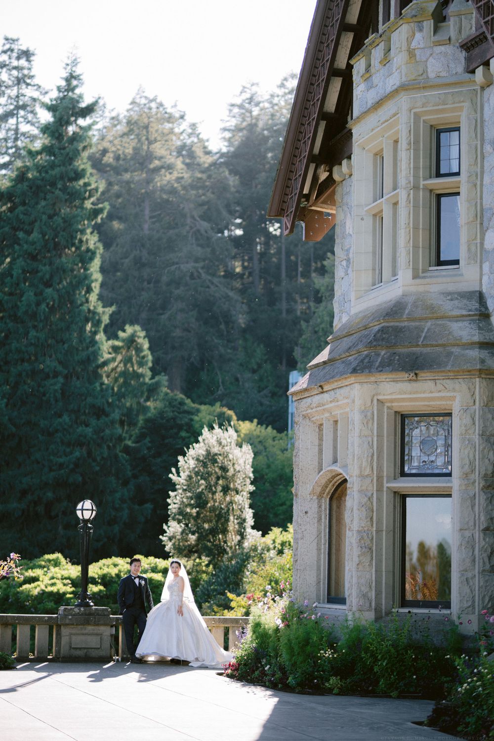 Bride in flowing gown stands near Victorian-style mansion with forest backdrop.