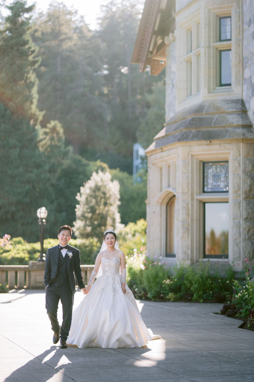Sunlit couple walks along pathway beside elegant stone mansion.