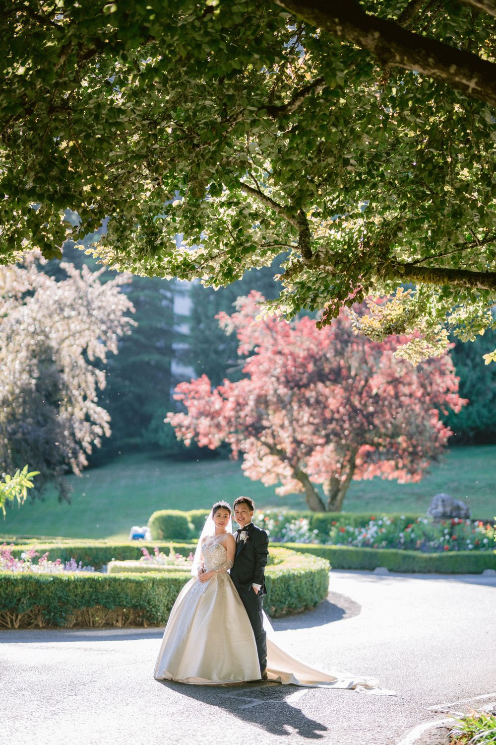 Couple poses in garden with cherry blossoms and lush greenery in background.