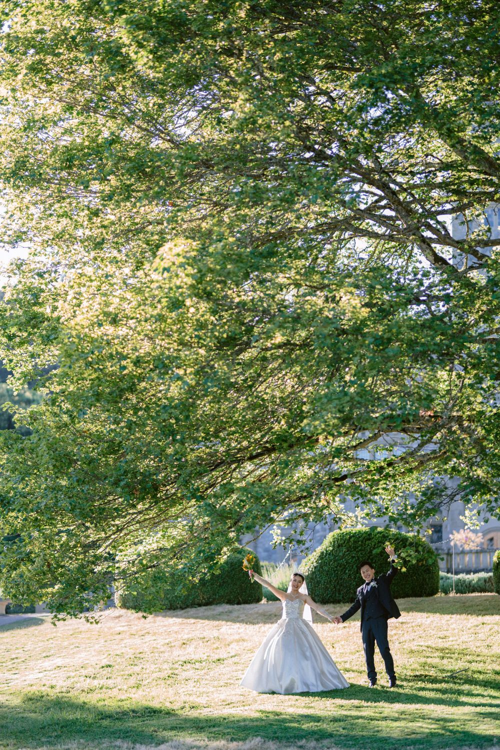 Newlyweds pose under sprawling tree branches in a sunlit garden setting.