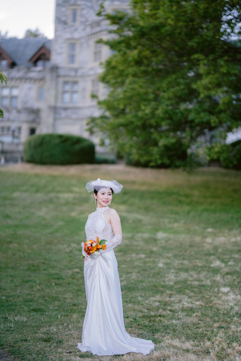 Elegant bride in white lace dress holds orange bouquet against stone castle backdrop on manicured lawn.