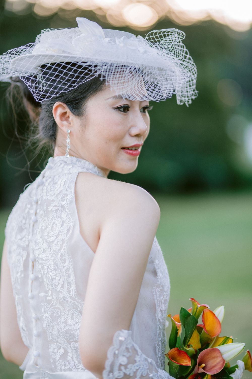 Bride in vintage-inspired lace dress and netted hat holding orange flowers in garden setting.