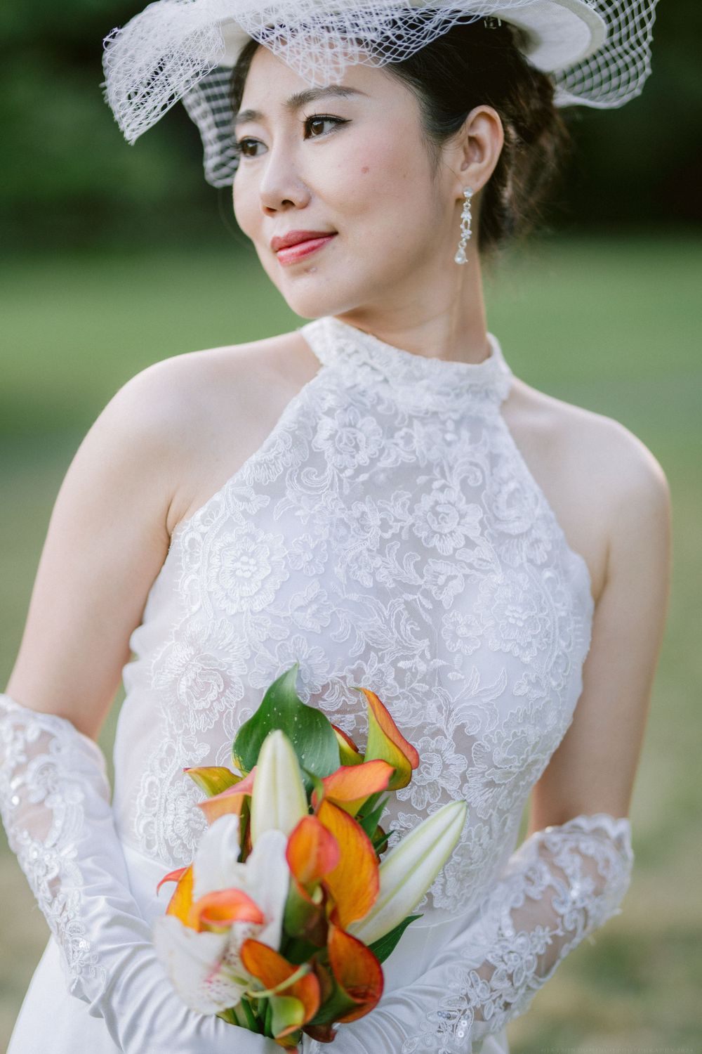 Bride in high-neck lace gown holding orange and white lily bouquet in garden setting.