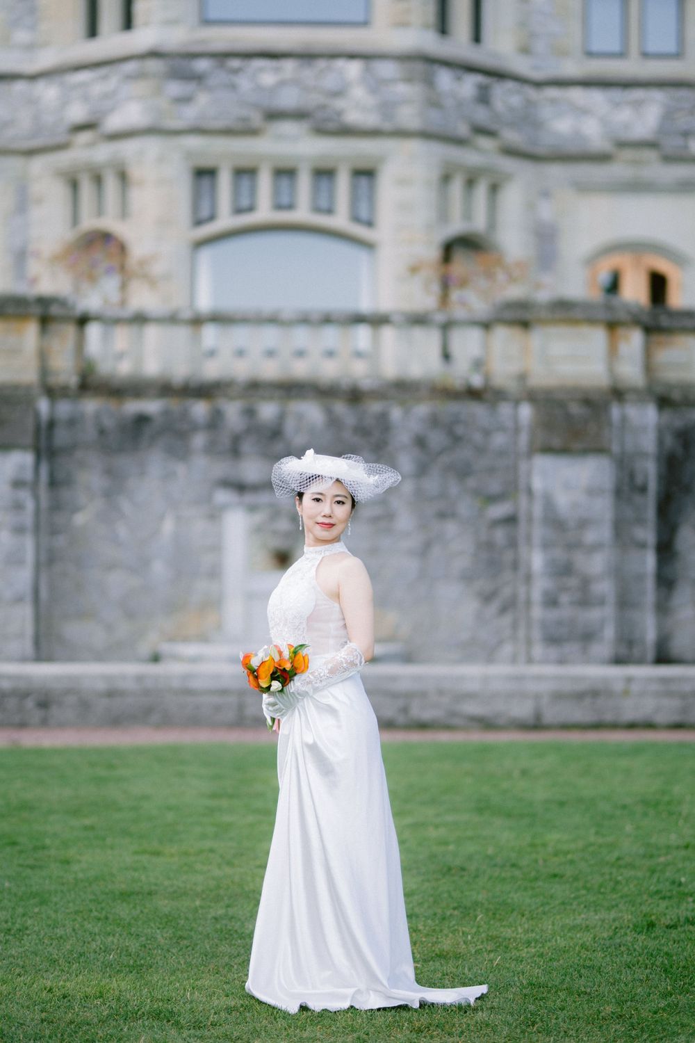 Elegant bride in white gown and ornate hat holds bouquet while standing on lawn in front of historic stone building.
