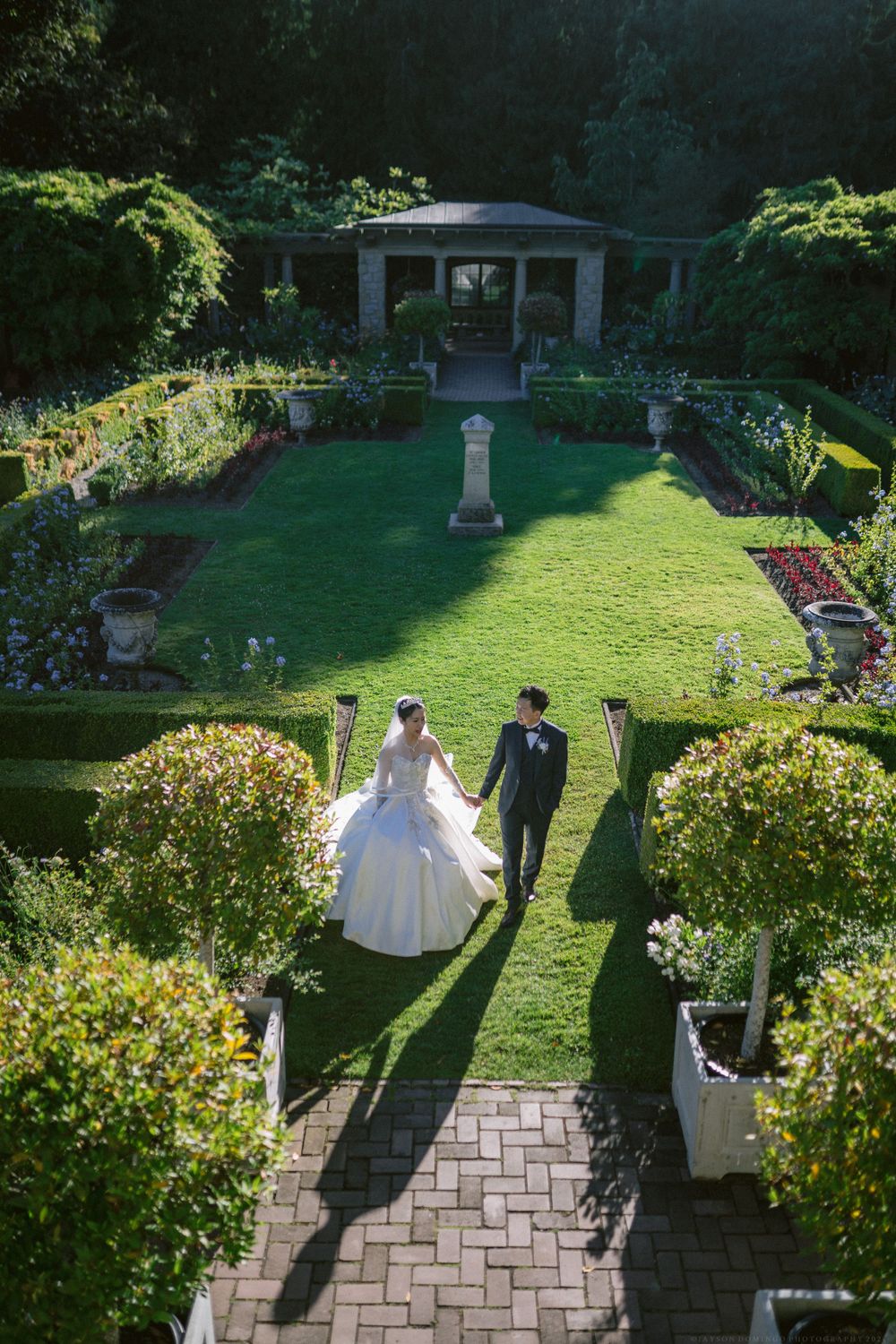 Aerial view of newlyweds walking through formal garden with gazebo and manicured hedges.