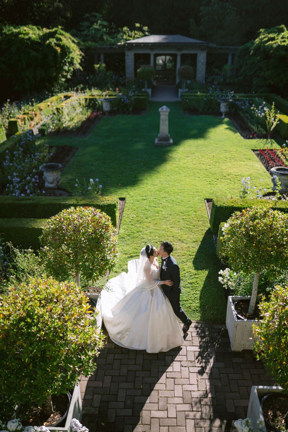 Bride and groom embrace in sunlit formal garden with gazebo and flowering borders.