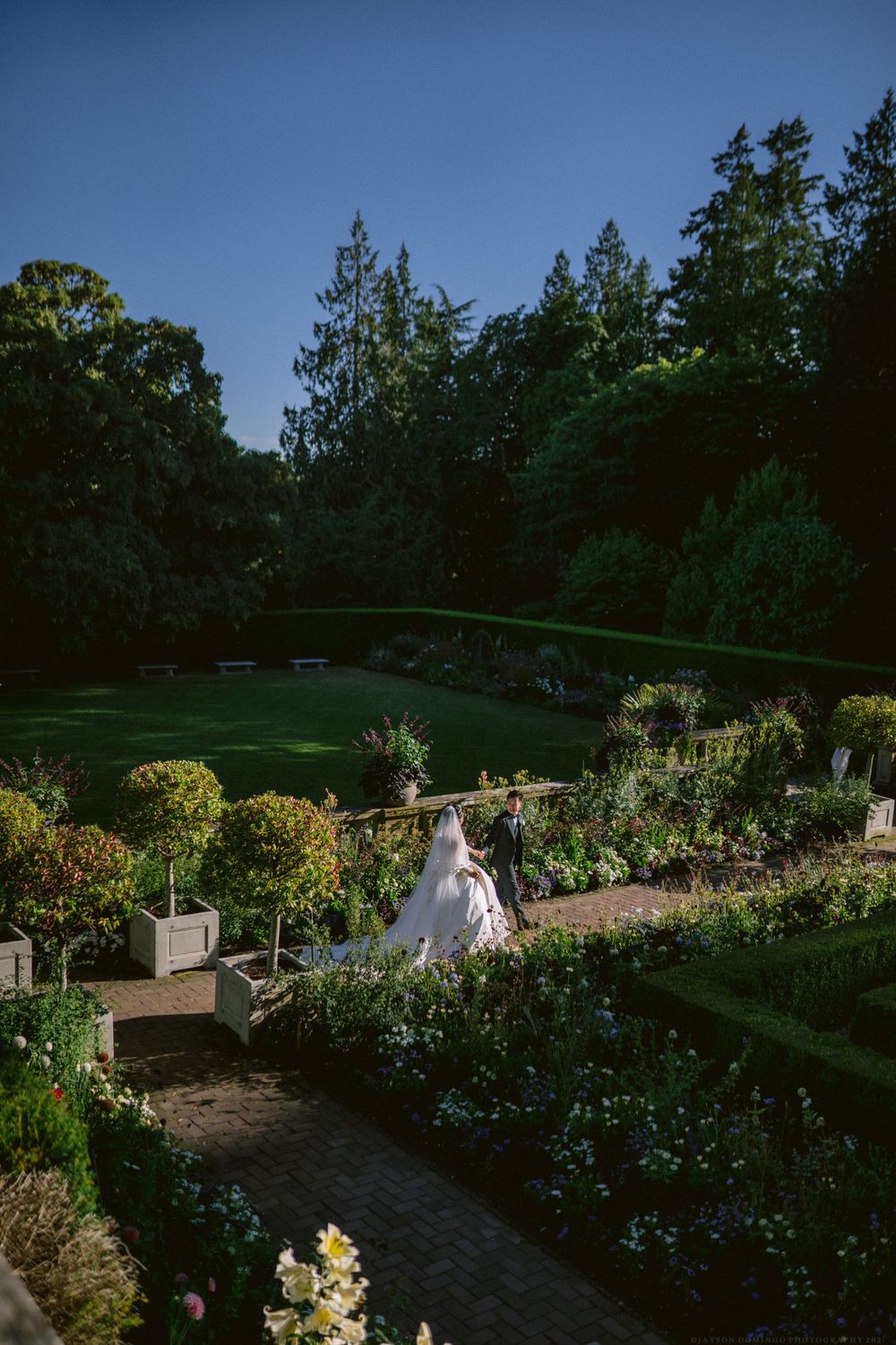 A couple in wedding attire walks through a beautiful garden path with blooming flowers.