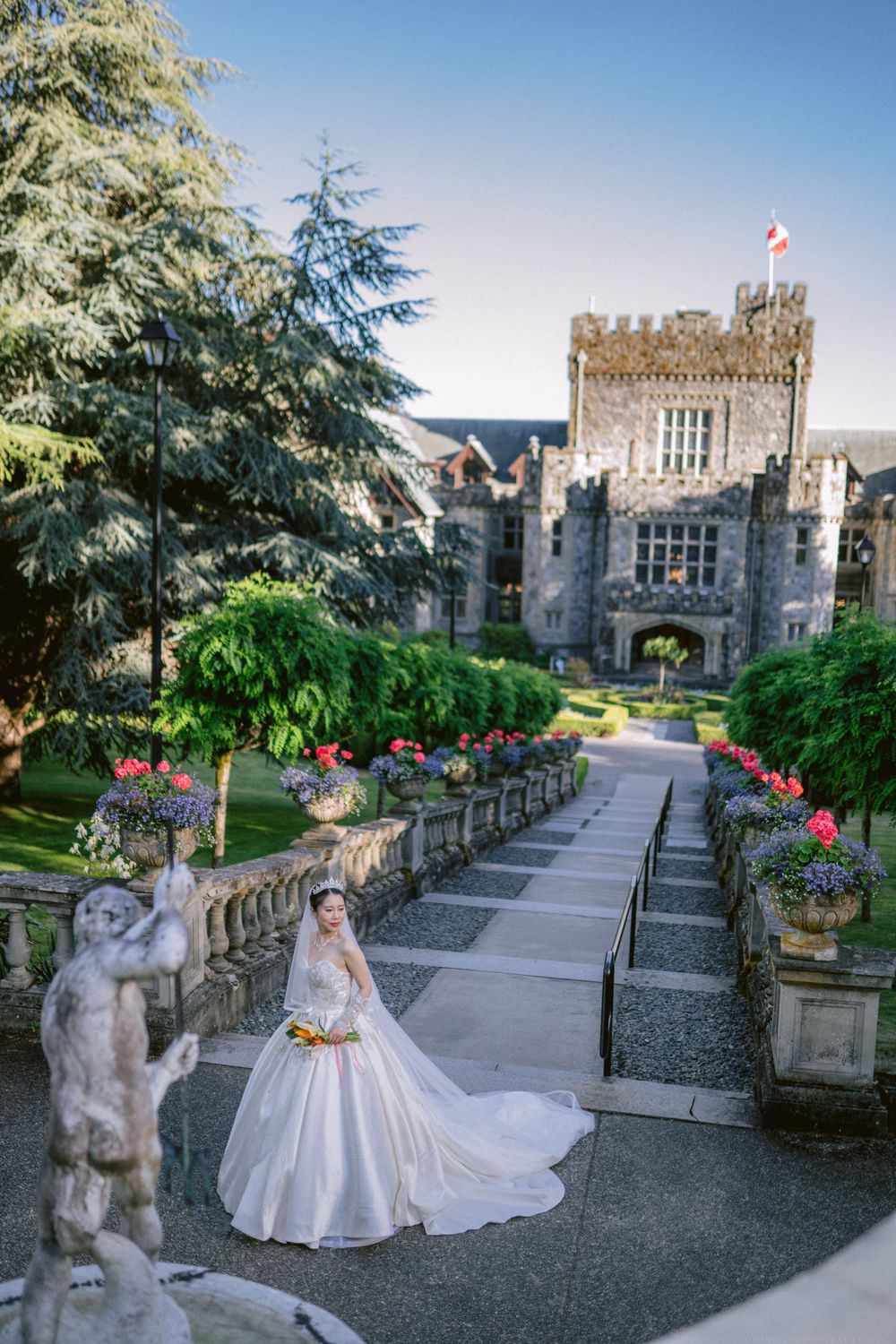 A bride in a flowing white gown stands before a majestic castle entrance with stone steps and flowers.