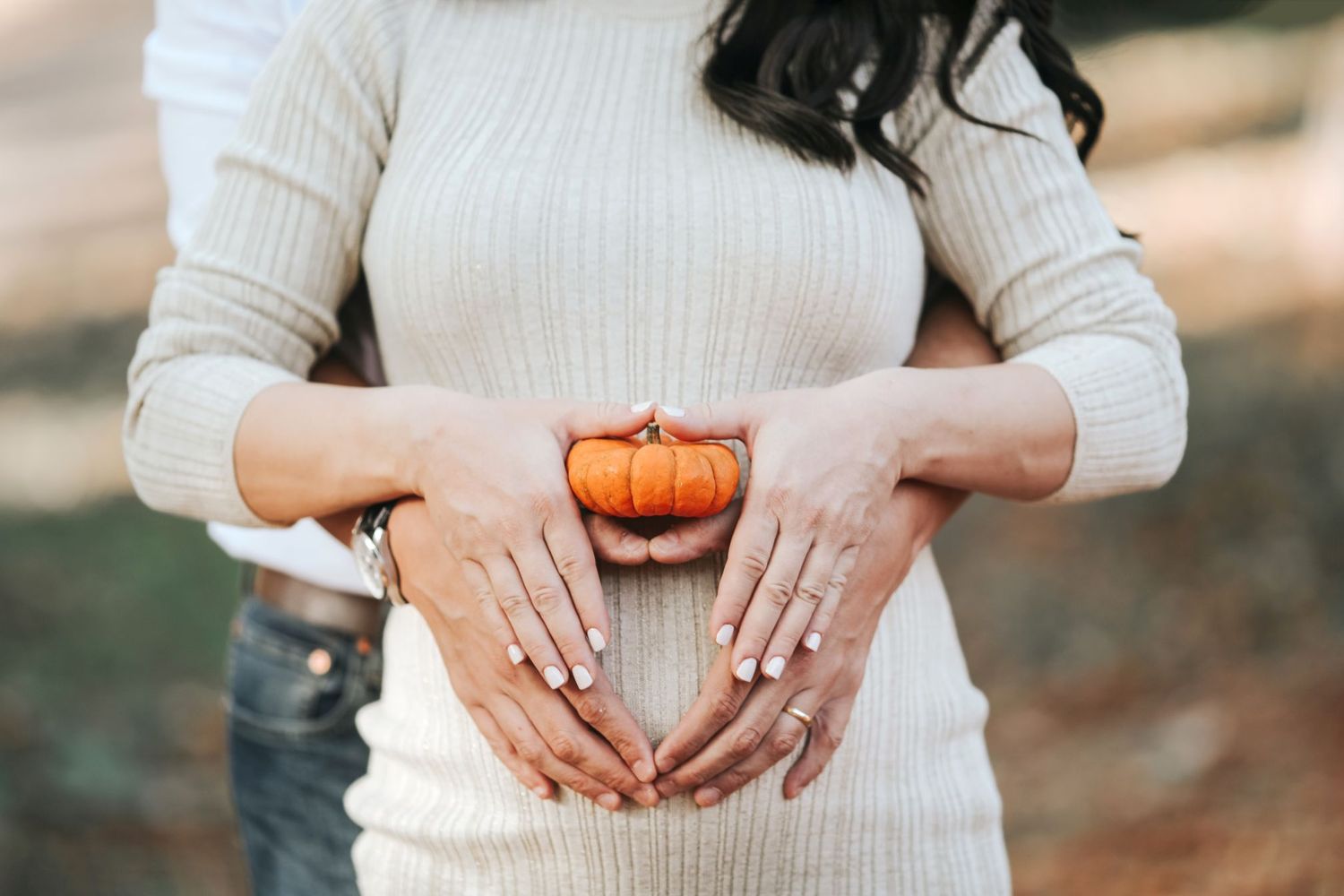 Hands form heart shape around small decorative pumpkin against white sweater.