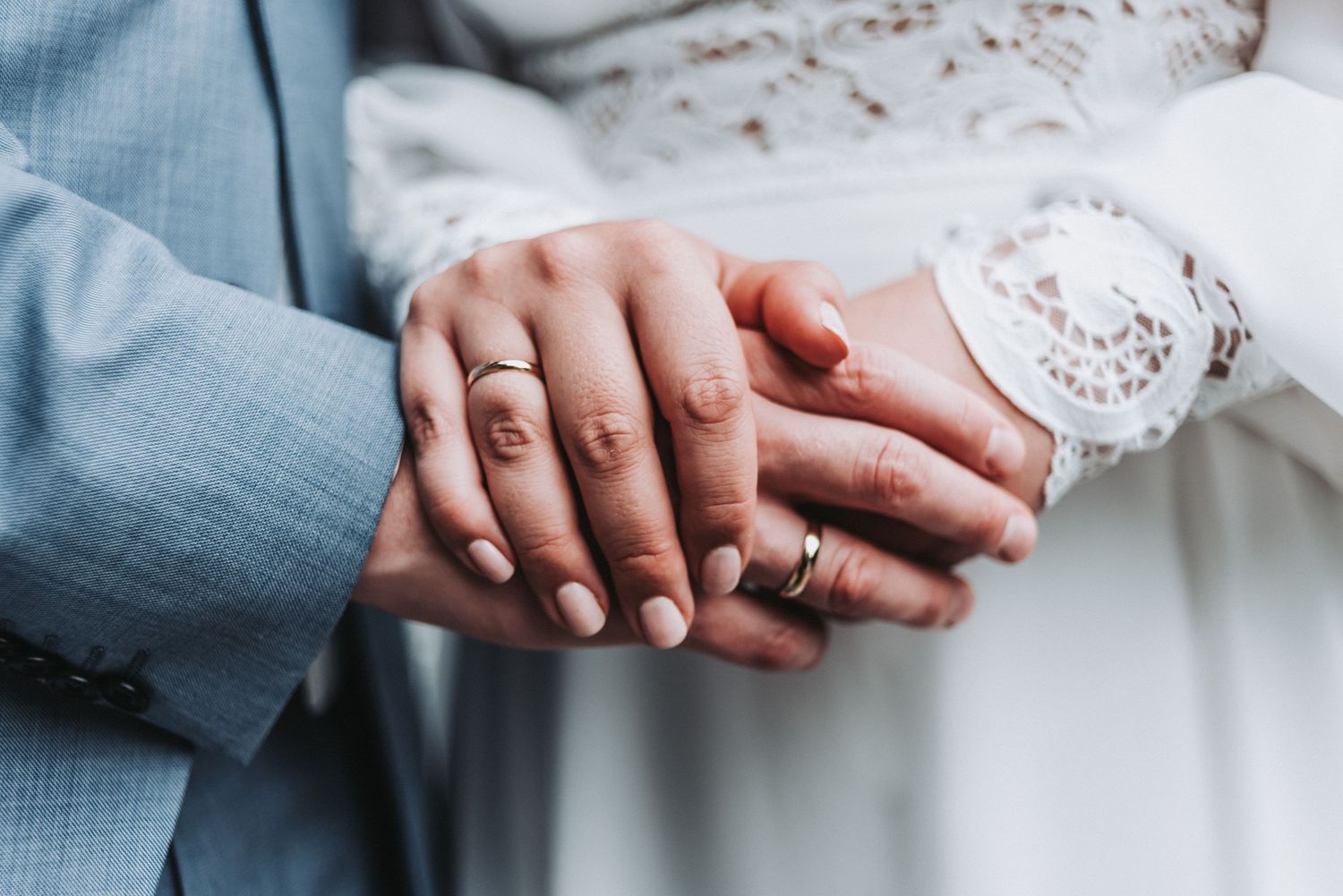 Close-up of newlywed couple's hands with wedding rings against light blue suit.