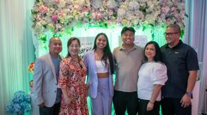 A group photo of party attendees standing together in front of a floral backdrop with blue and green lighting.