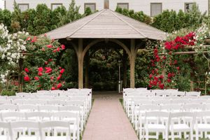 Outdoor wedding ceremony setup with white chairs and wooden gazebo decorated with red climbing roses.