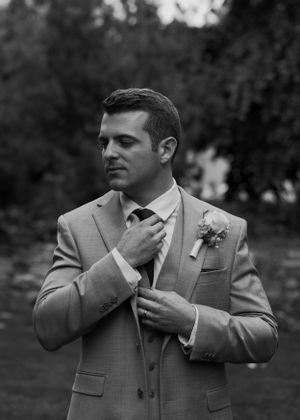 Groom adjusts his tie while wearing a light gray suit with boutonniere in black and white portrait.