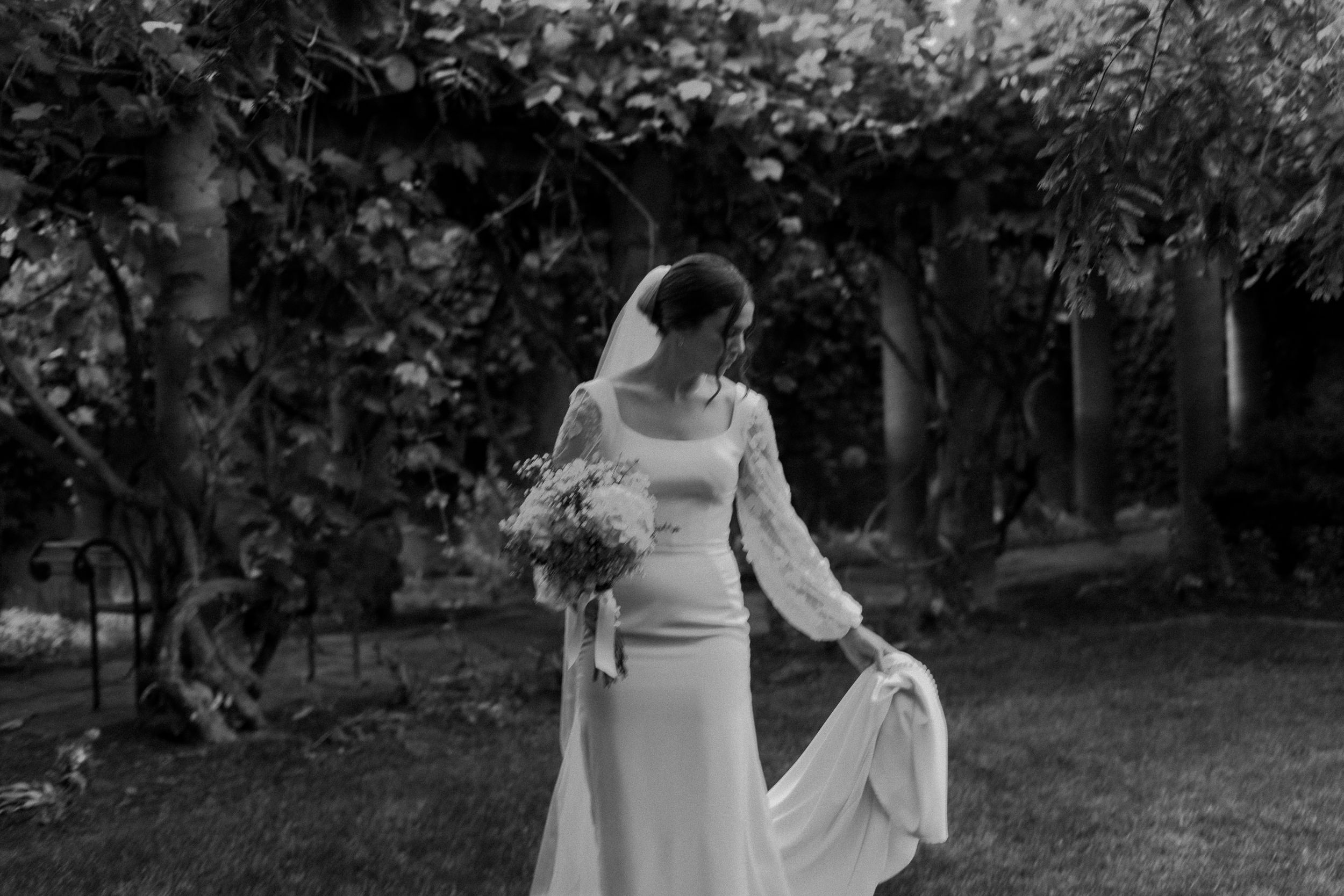 Black and white portrait of bride in long sleeve gown holding bouquet in garden setting.