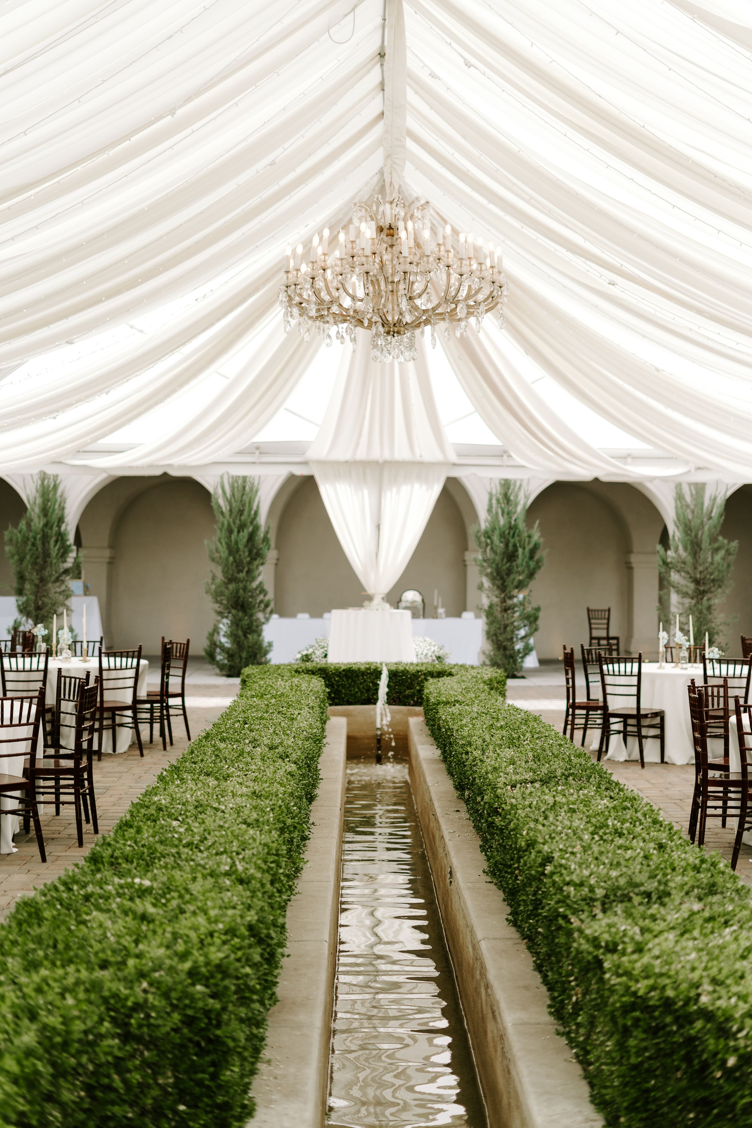 Elegant tented reception space with chandelier and boxwood-lined water feature creating central aisle.