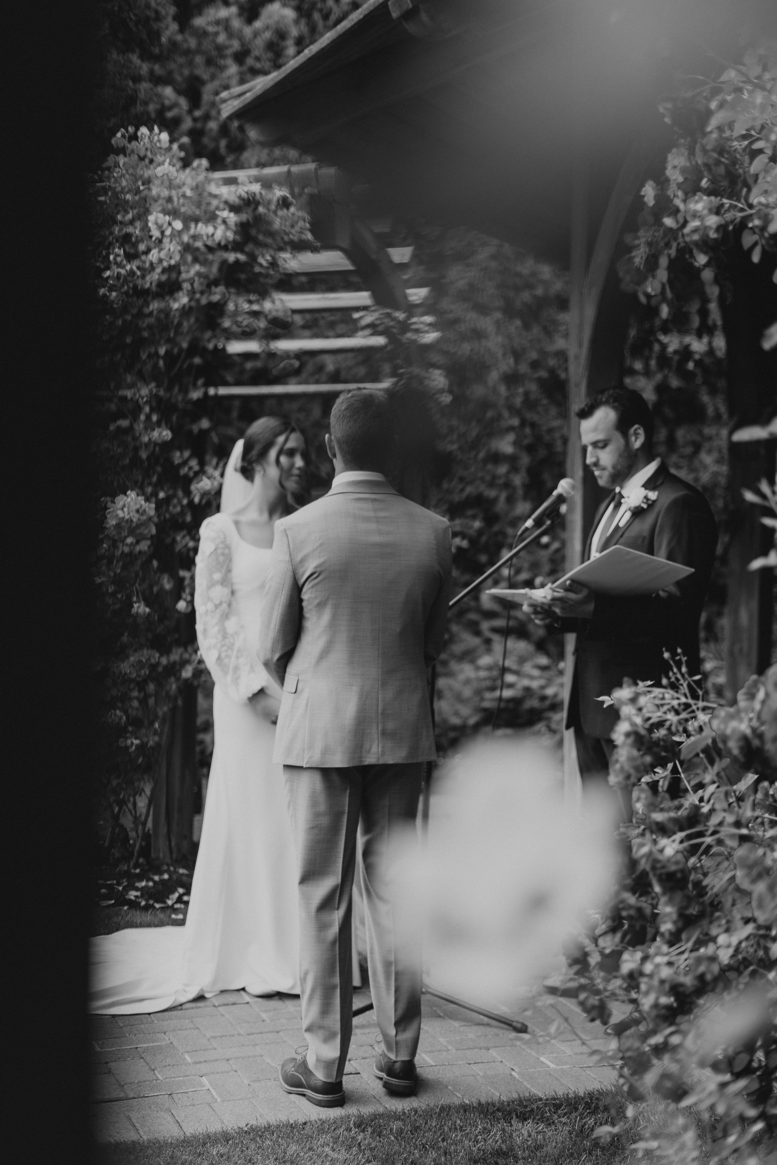 Black and white photo of a wedding ceremony under a garden gazebo with flowering vines.