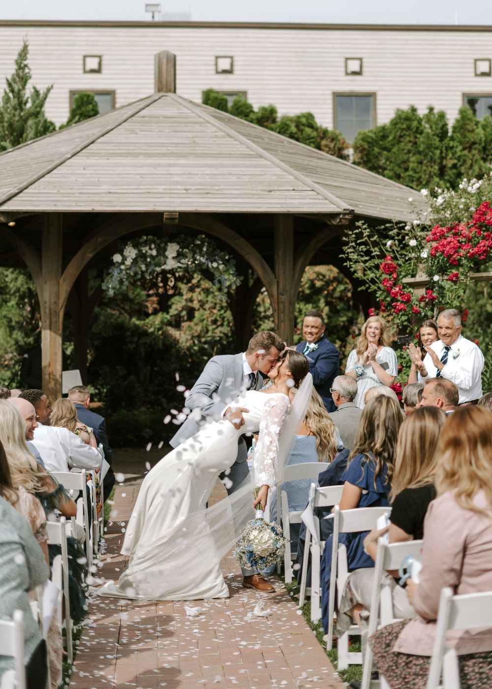 Wedding ceremony under a wooden gazebo decorated with flowers and guests seated on white chairs.