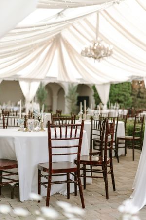 Elegant wedding reception setup with mahogany chiavari chairs and white linens under draped tent.
