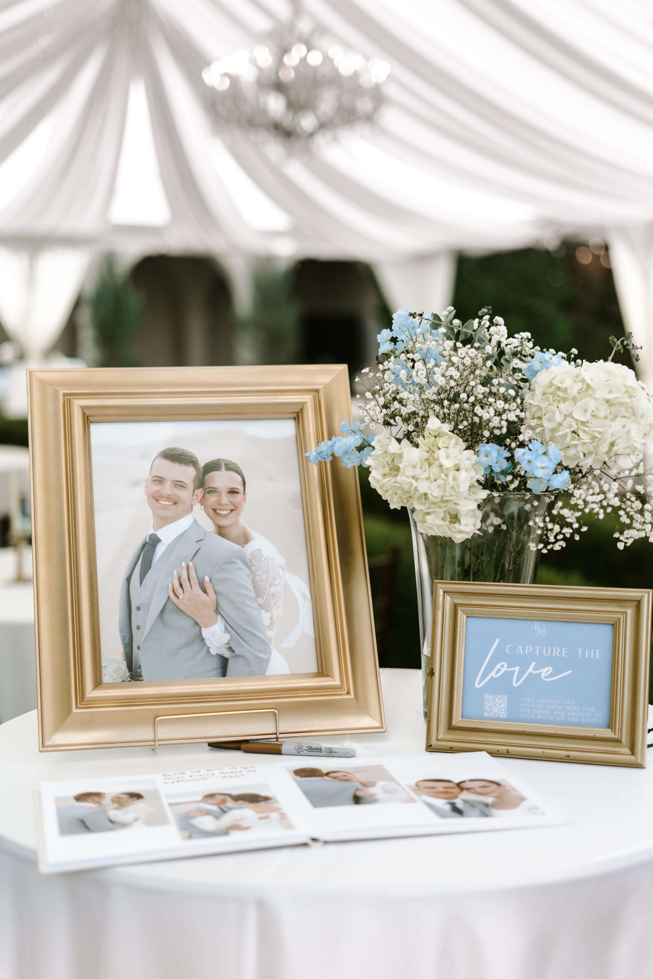 Wedding reception table with framed photos and blue and white floral arrangements under white tent.