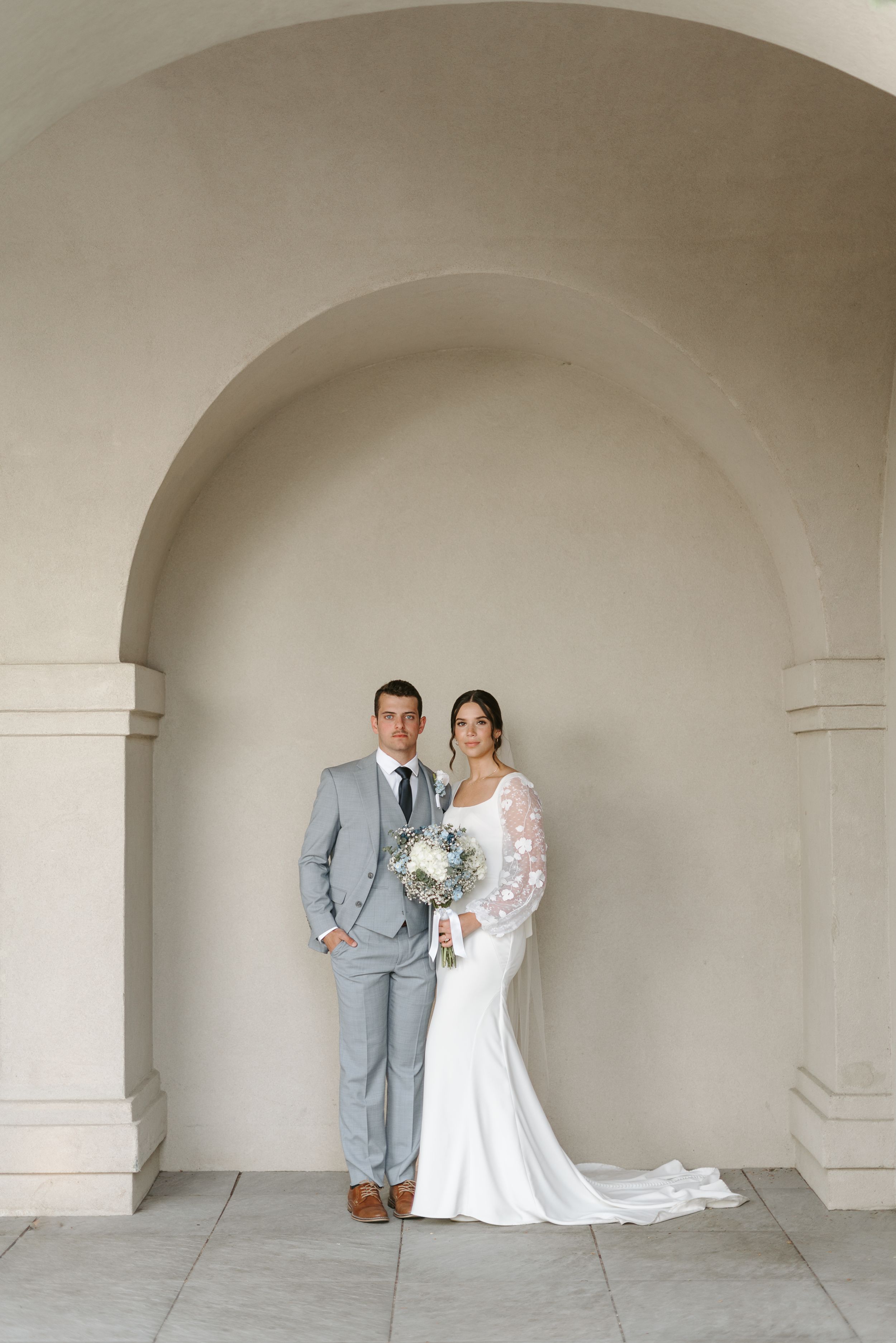 Wedding couple poses under architectural archway with bride in lace gown and groom in light gray suit.