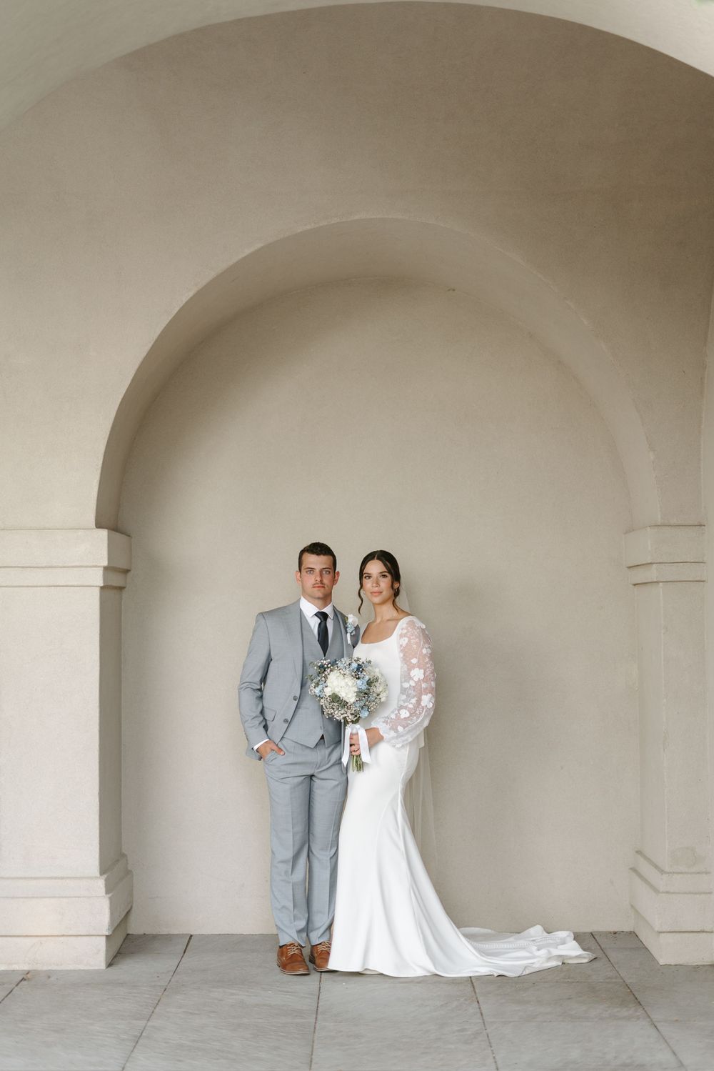 Wedding couple poses under architectural archway with bride in lace gown and groom in light gray suit.
