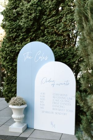 Modern light blue and white arch-shaped wedding signage displaying order of events against greenery.