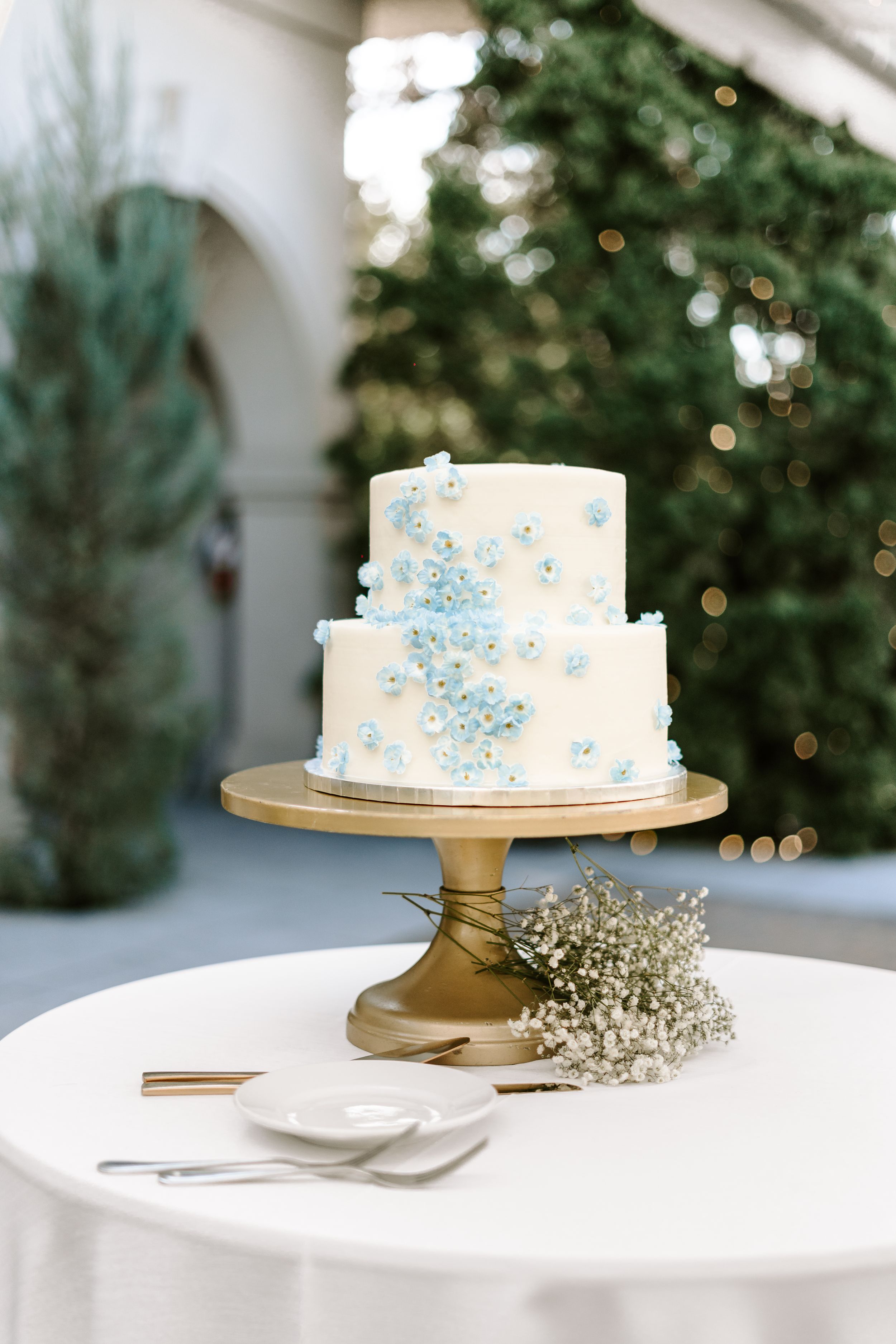 Two-tiered white wedding cake decorated with light blue pearls on gold stand with baby's breath accent.