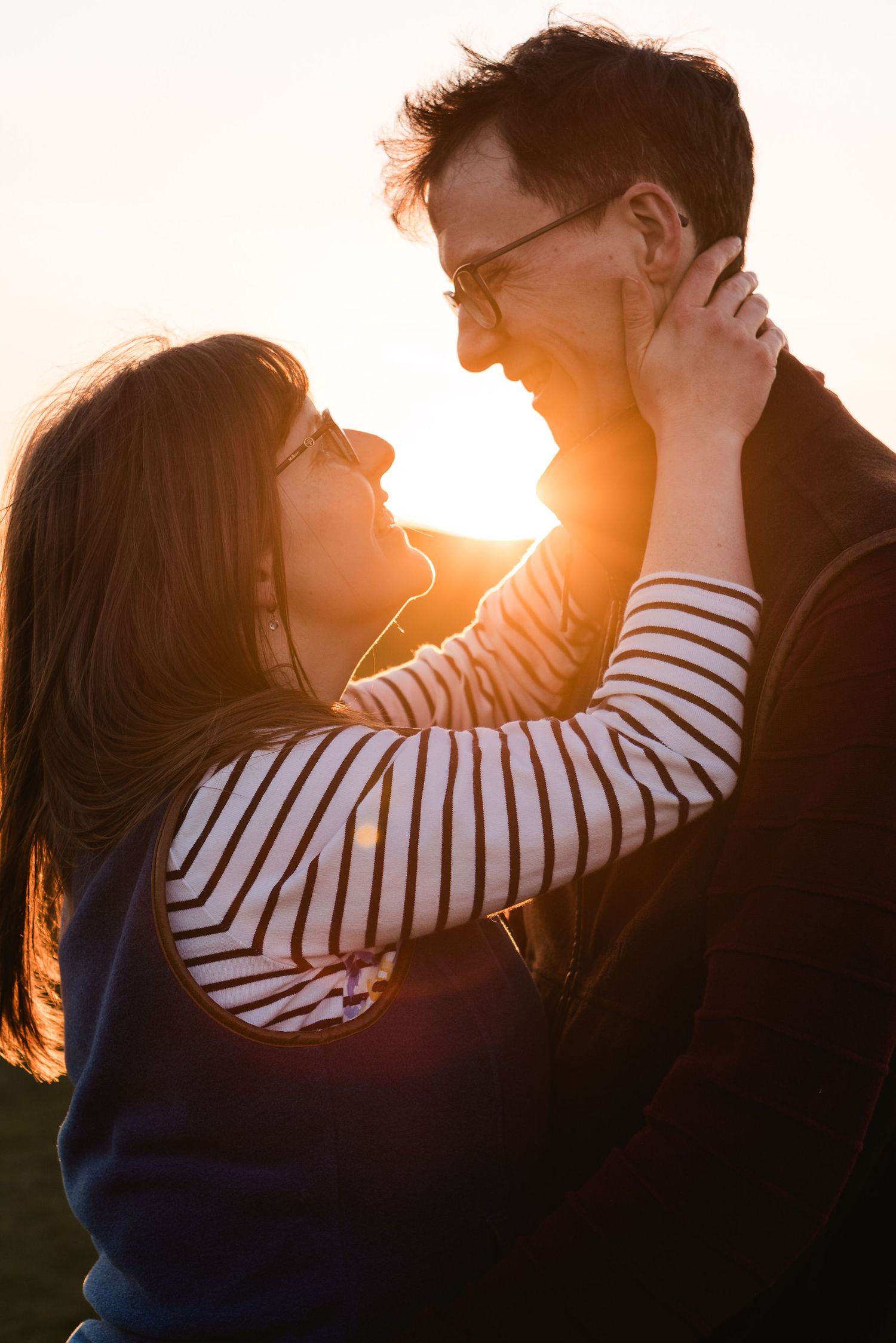 A young couple looking lovingly at each other, she has her hands on the mans face. The sun is shining through their faces, at goldenhour