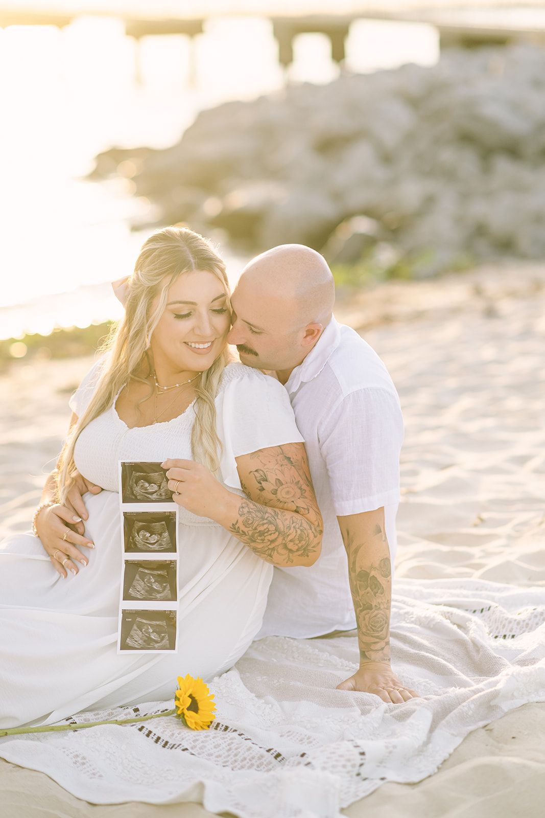 Couple sitting on beach blanket sharing ultrasound photos during sunset.