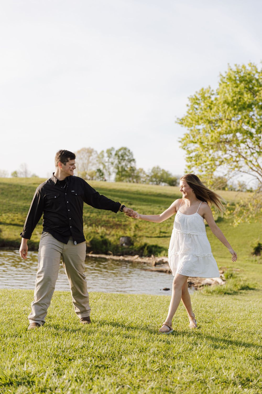 A couple dances together on green grass near a pond on a sunny spring day, casting playful shadows.