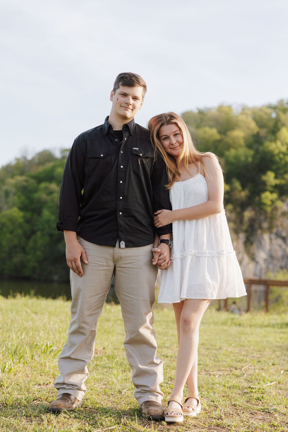 A couple in coordinating casual outfits stands together in a grassy field on a sunny day with trees in the background.