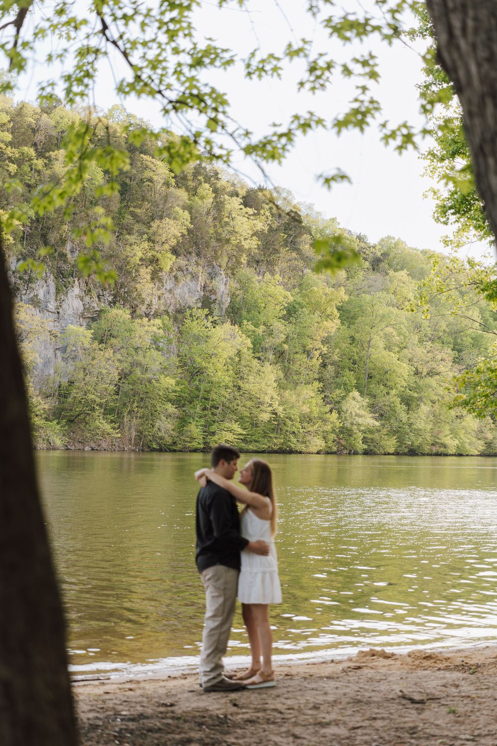 A couple embraces by a tranquil lake surrounded by lush green trees and rocky cliffs on a peaceful summer day.