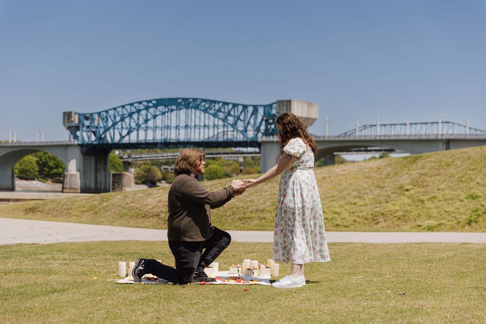 A romantic proposal scene takes place on a grassy hill with a blue bridge in the background on a sunny day.