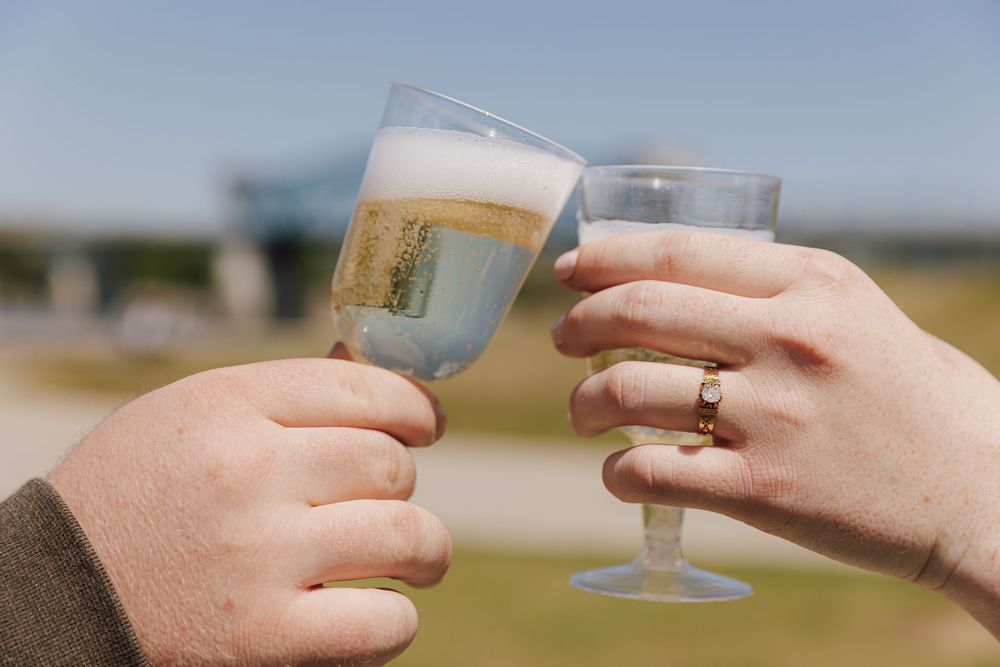 Two hands toast with champagne glasses outdoors, one wearing a gold wedding ring.