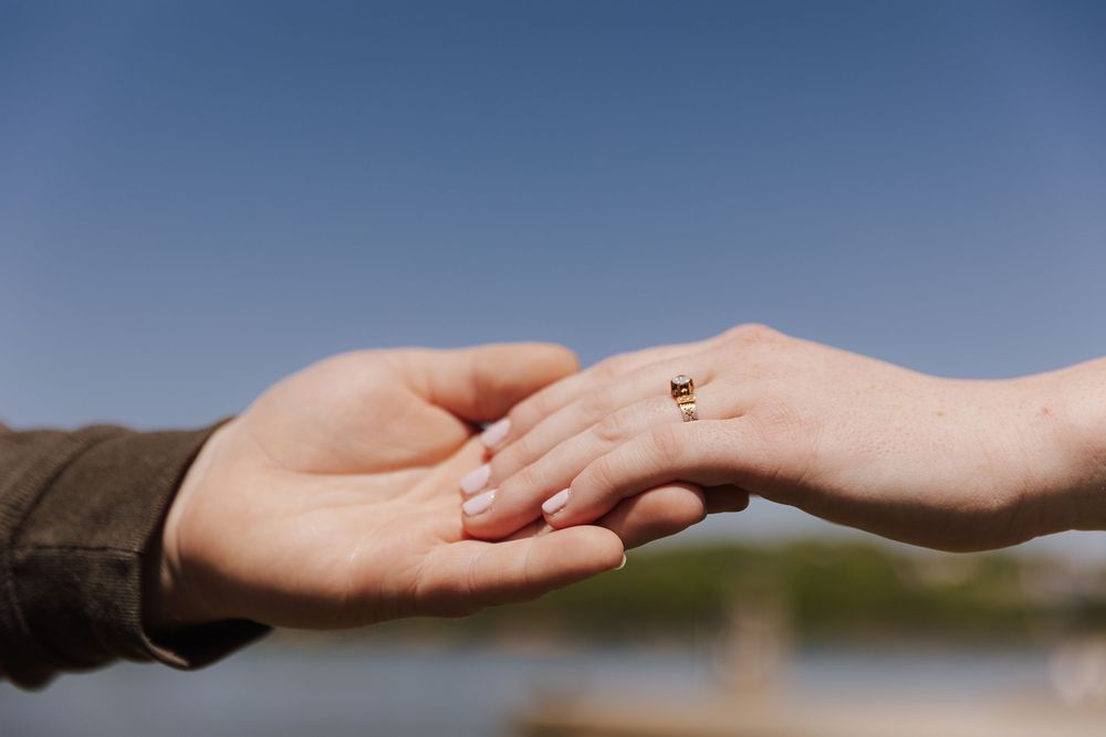Two hands gently touch in a tender moment with a diamond ring visible against a blurred outdoor backdrop.