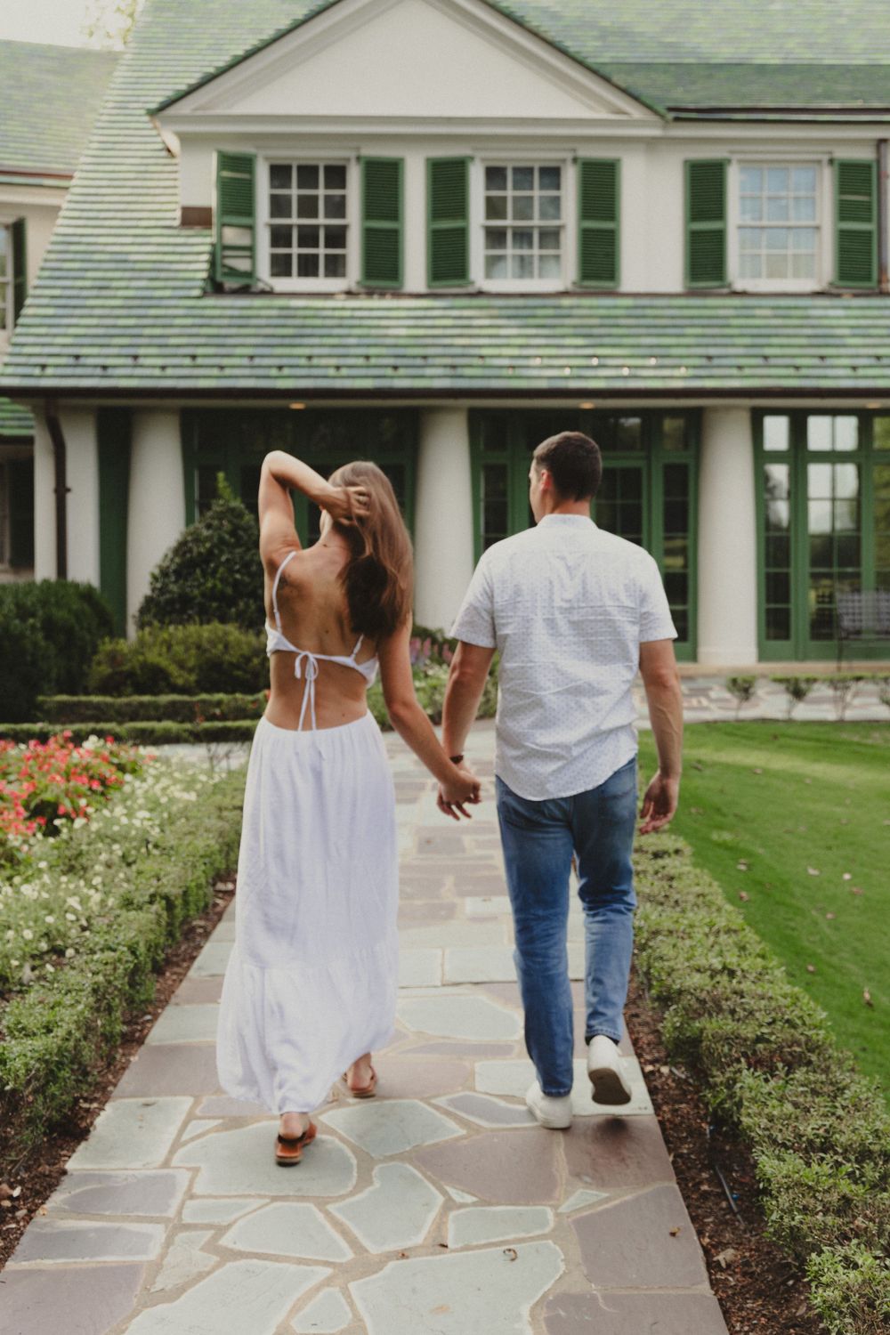 Walking along stone path in front of classic house with green shutters and flower garden.