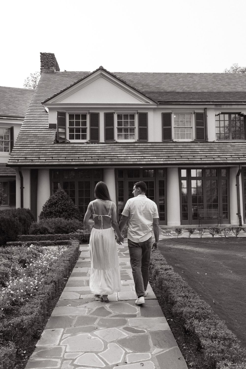 Black and white photo of couple walking hand in hand towards colonial style house.