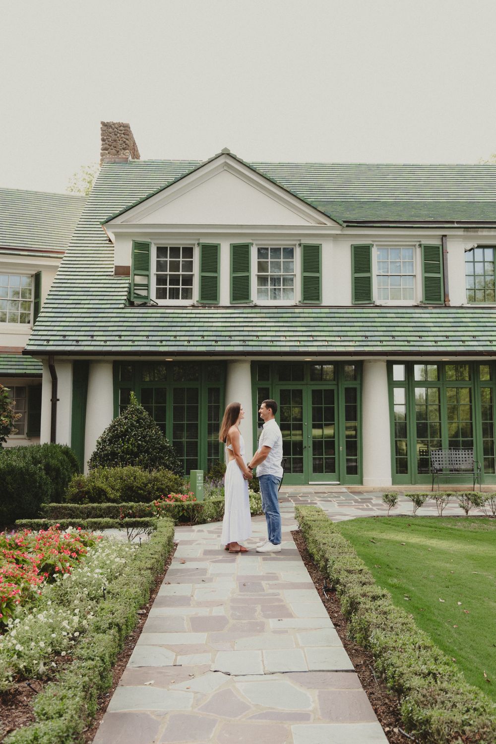 A couple shares an intimate moment on a stone path leading to a stately white house with green shutters and roof.
