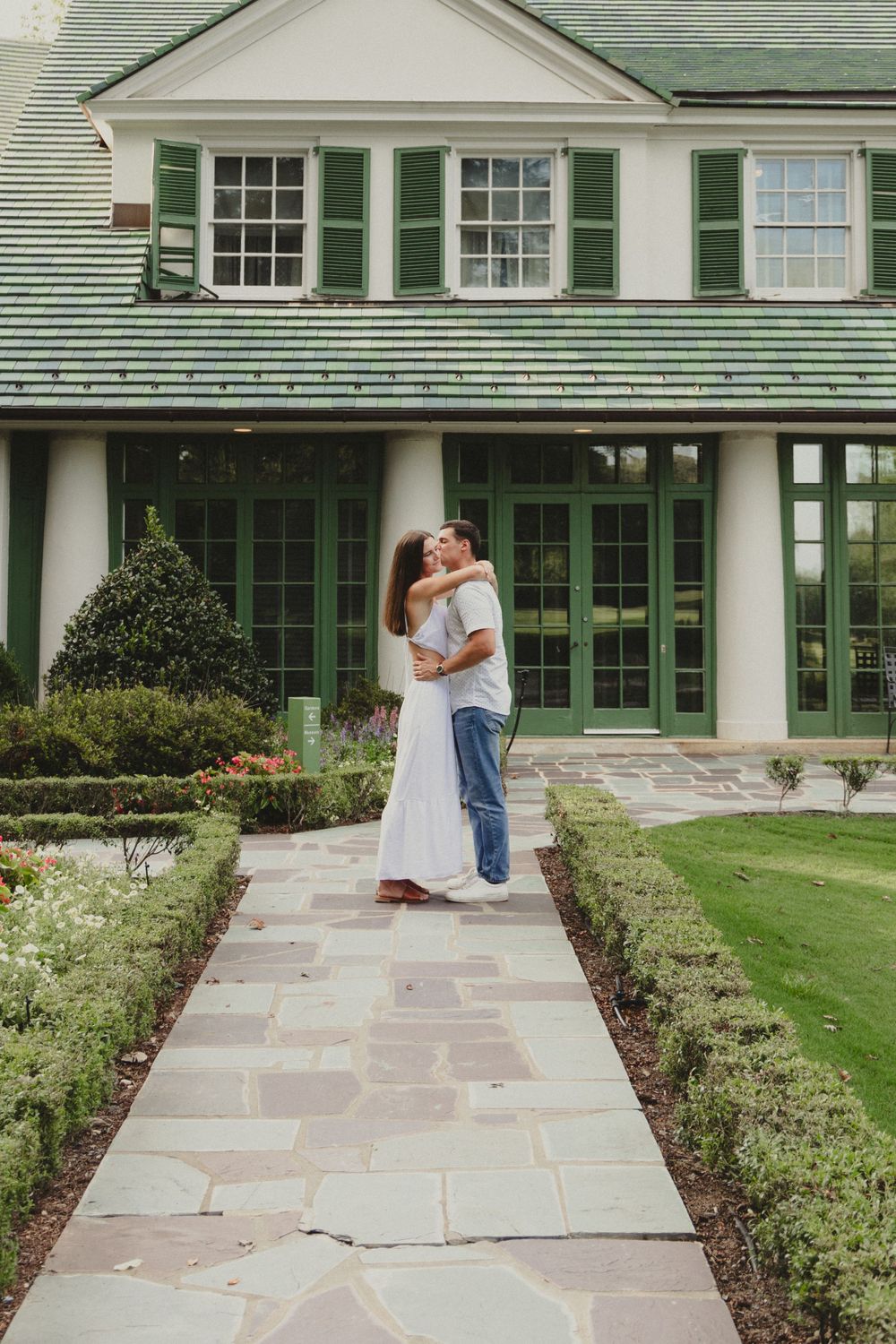 A romantic moment between two people in front of a colonial-style home with manicured gardens.