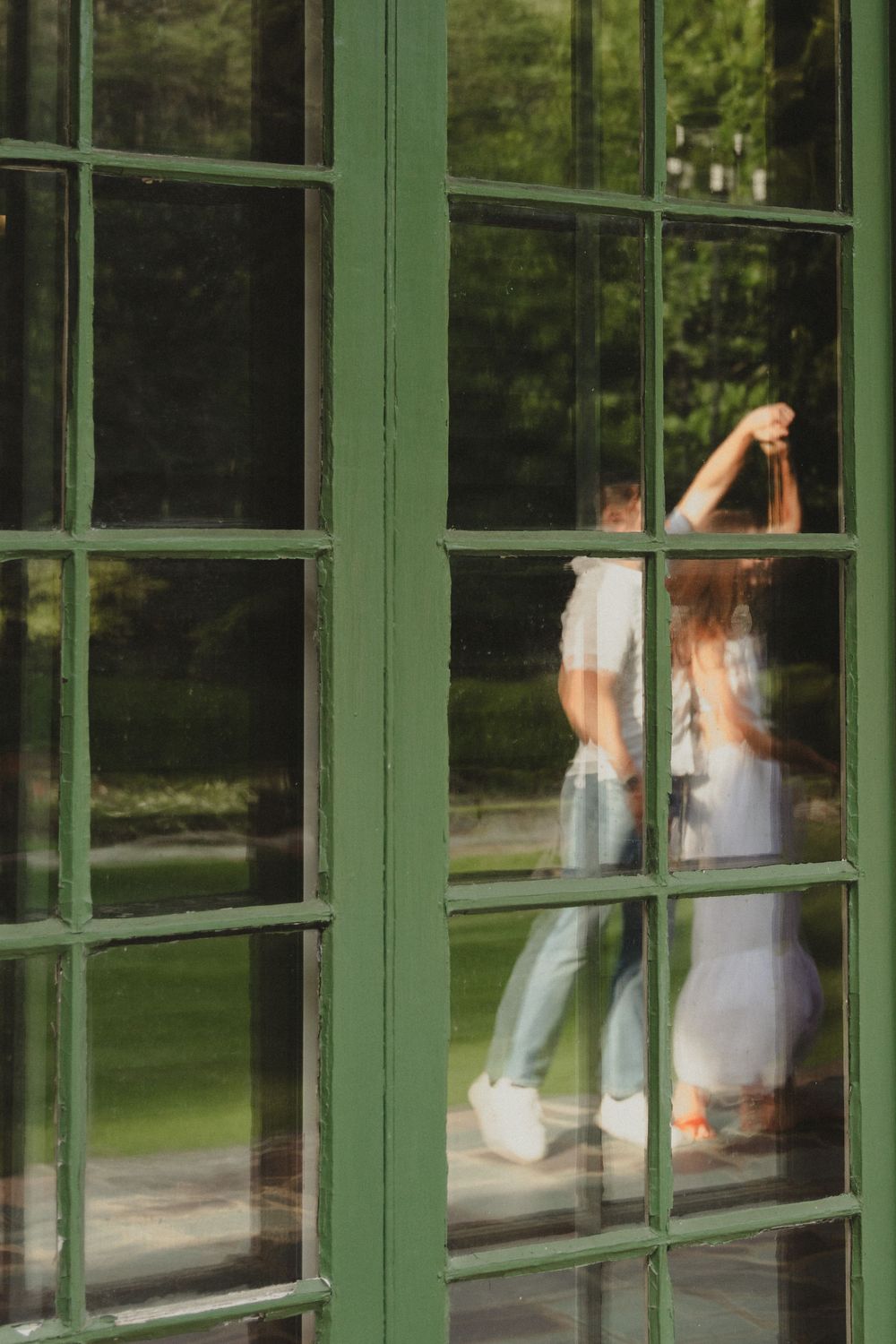 A reflection through green-framed windows shows two people dancing inside the building.