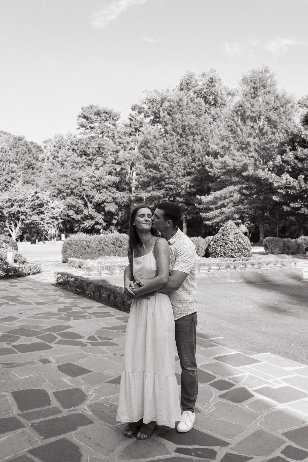 A black and white photograph of two people embracing on a stone patio surrounded by trees.