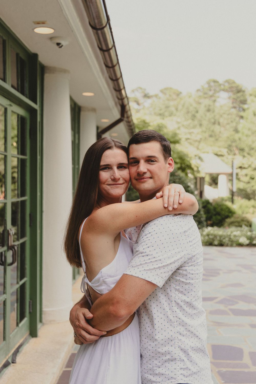 A couple embraces against a white colonial building with green trim and windows.