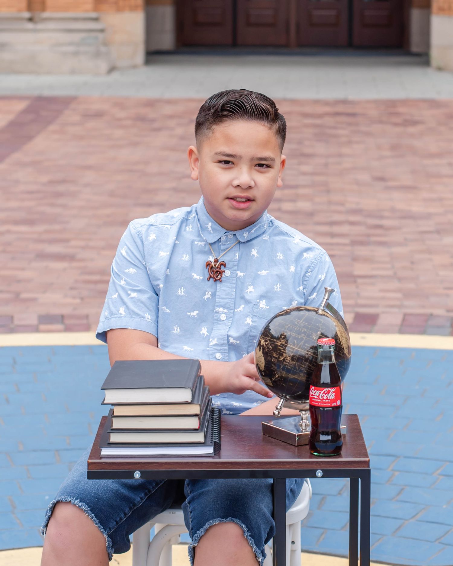 Student in patterned shirt sits at outdoor table with books and refreshments.